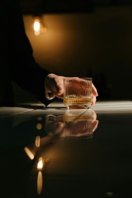 Close-up of a hand holding a whiskey glass with reflections on a bar counter in a dim light.
